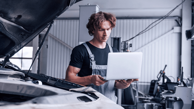 Mechanic holding a laptop beside a car with an open bonnet.
